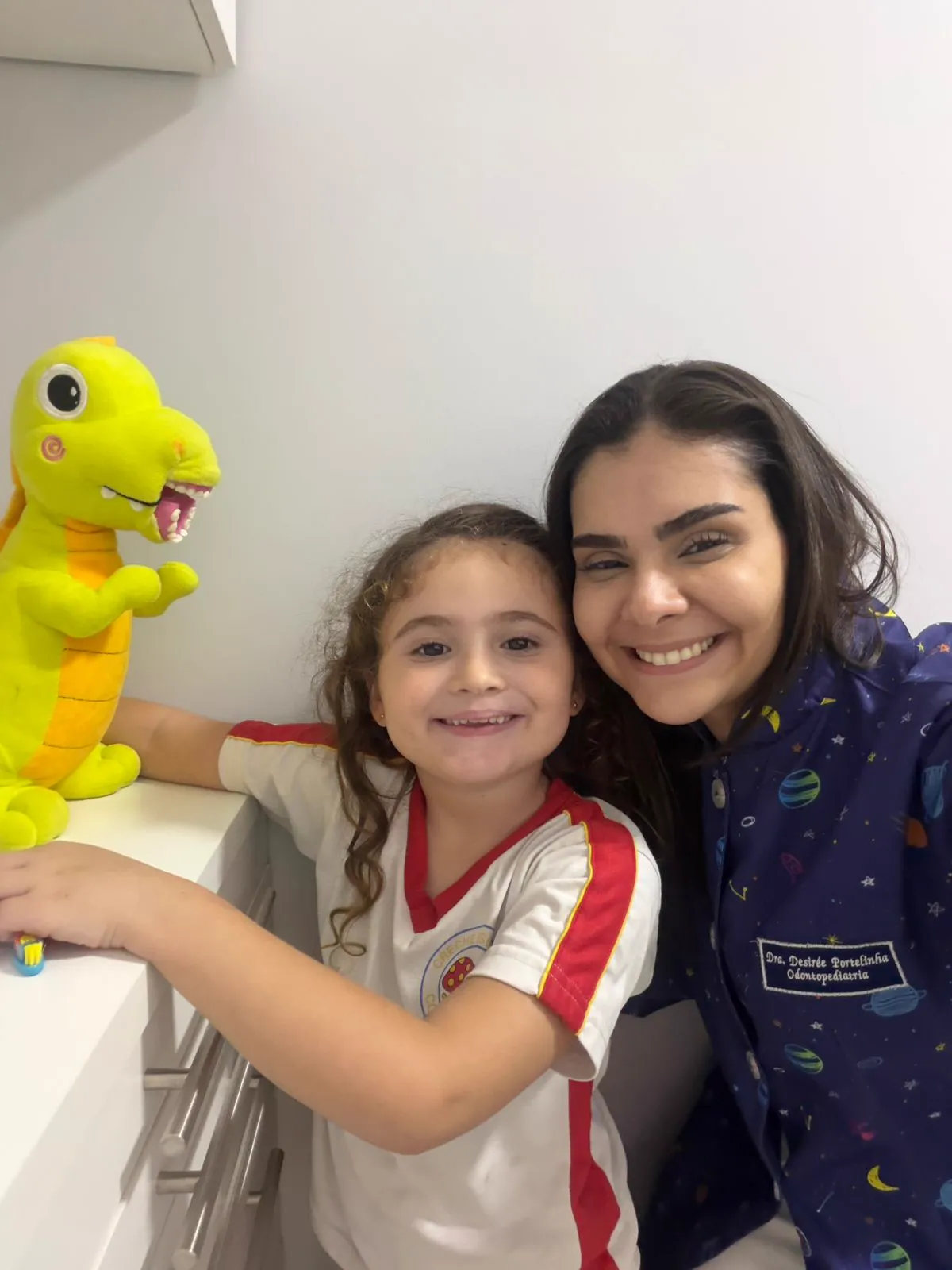 A Dra. Desirée e uma menina ao lado do Dino, a pelúcia de dinossauro amarela com dentes.